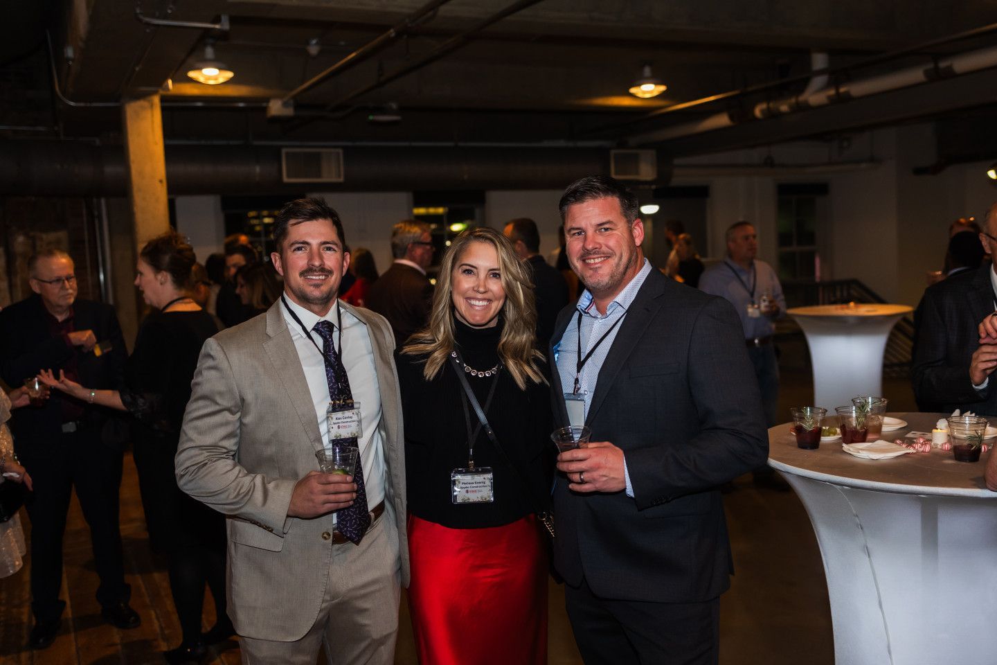 Three people smiling at a networking event, holding drinks. Woman in red skirt and black top, men in suits.