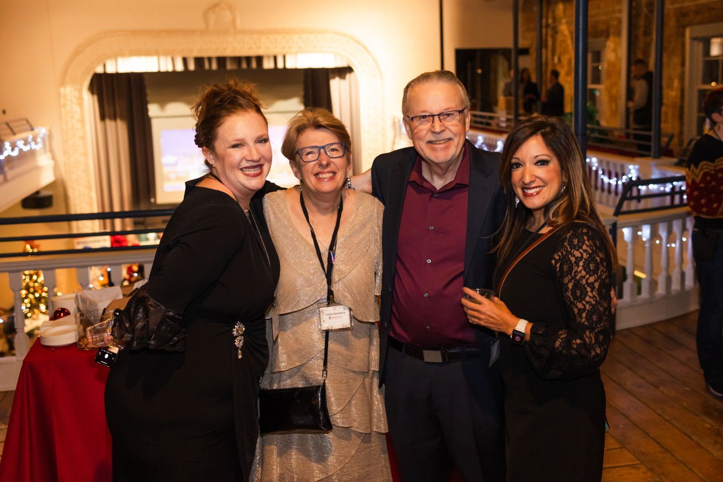 Four people smiling and posing together inside a building with a decorated railing, festive lighting.
