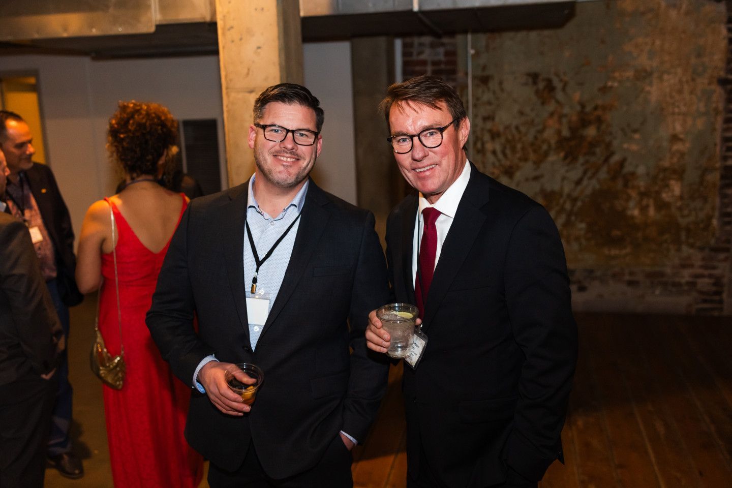 Two men in suits, wearing glasses, at an event. One holds a drink, smiling. Brick wall background.