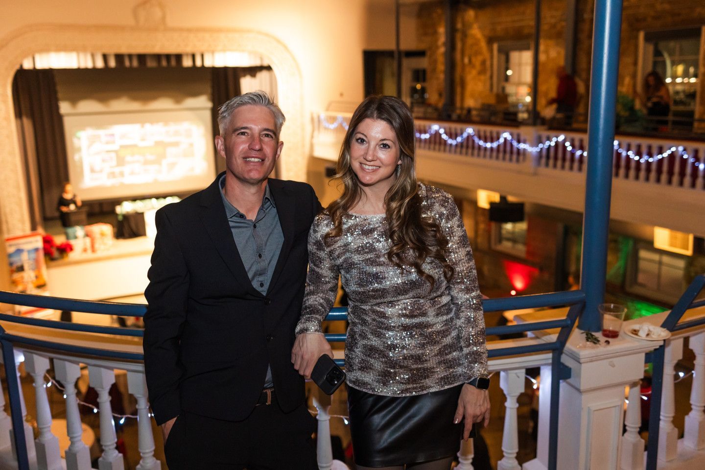 Man and woman pose on balcony, interior setting. Man in black suit, woman in sequined top and skirt.
