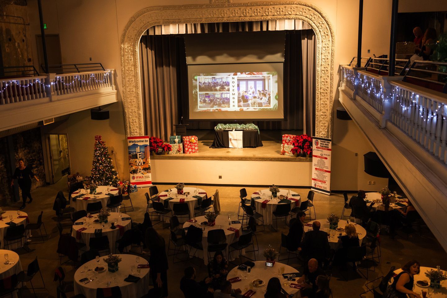 Audience seated at tables in a decorated auditorium. Stage with screen, gifts, and banners. Christmas tree on the left.