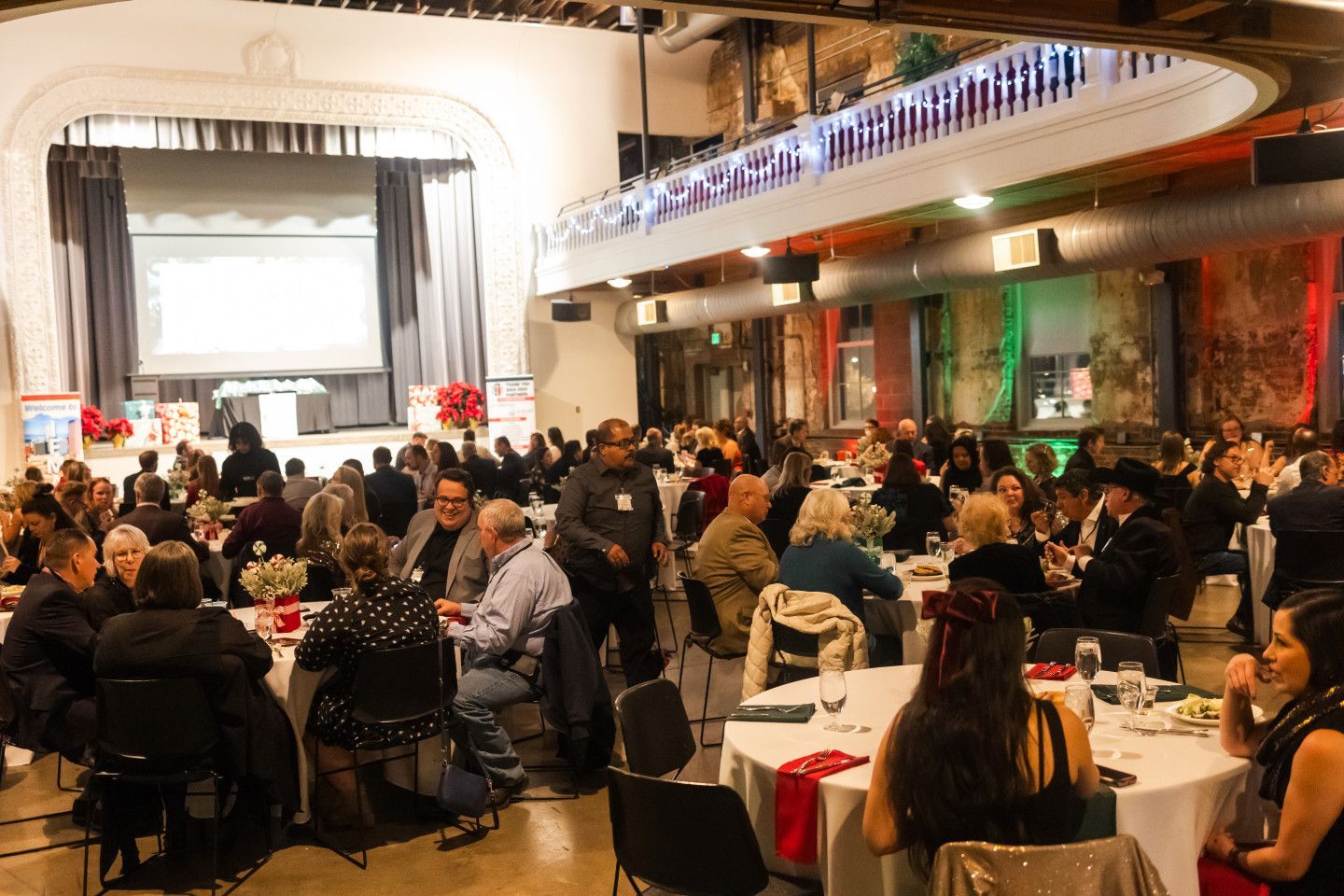 Large event: people at tables in a decorated hall. Stage with screen in the background. Festive atmosphere.