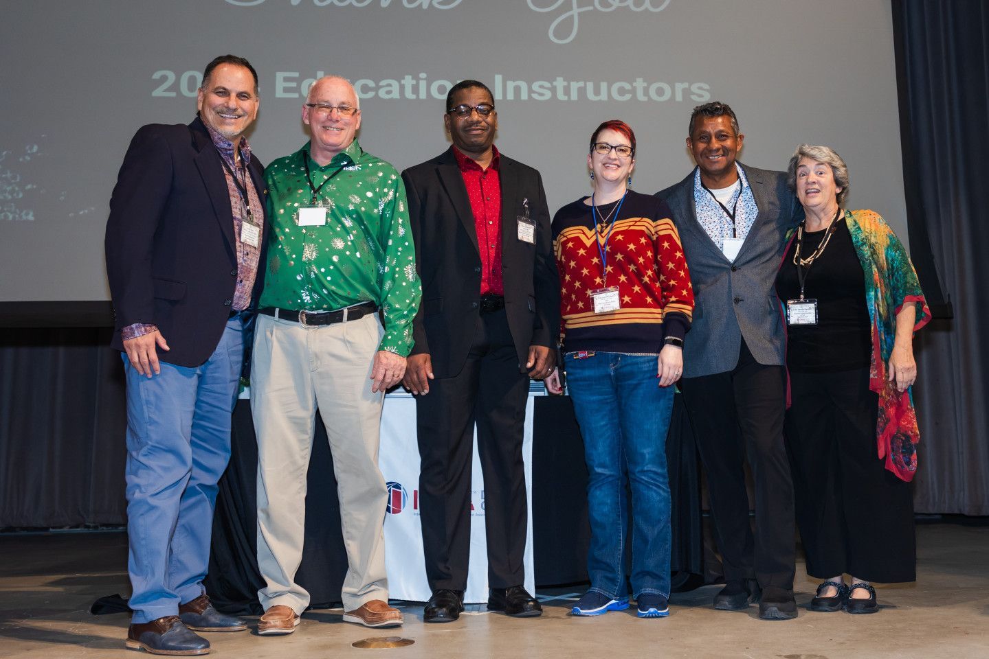 Group of six people posing for a photo on a stage with a screen in the background; they appear to be award recipients.