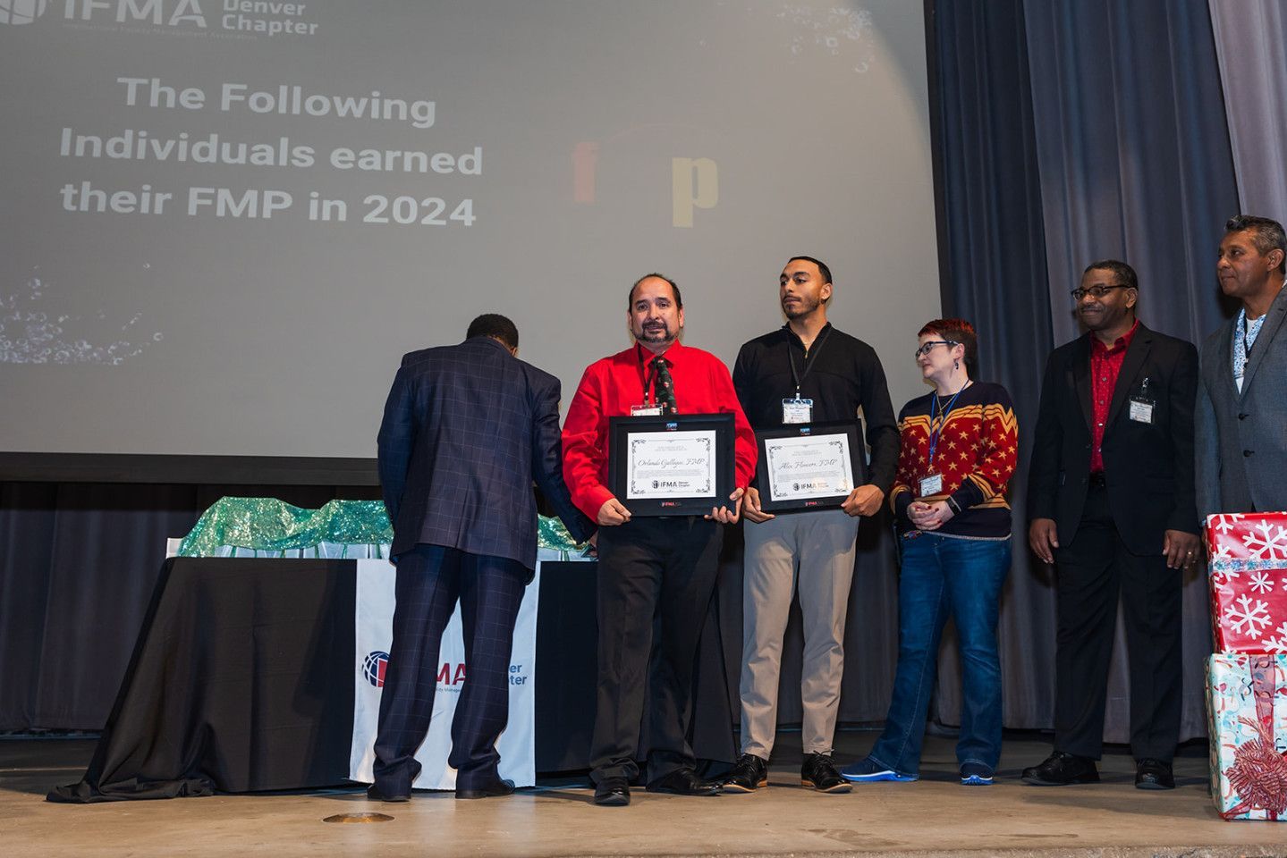People on stage holding certificates, in front of a screen displaying text. Award ceremony.