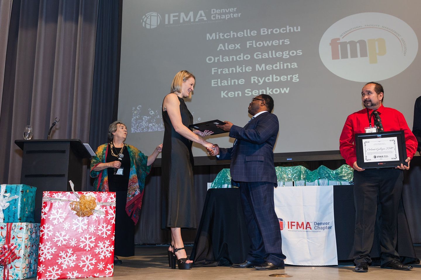 Woman receiving award from man on stage; other people and gifts in the background.