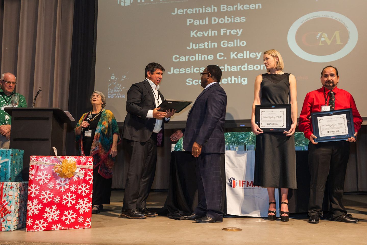 Awards ceremony: Man presents certificate to suited recipient. Woman and man hold certificates, gifts nearby.