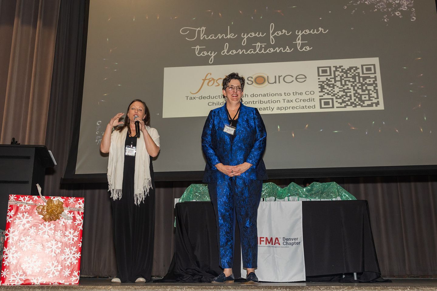 Two women on stage; one speaks into a microphone, the other stands. A backdrop shows a sponsor's logo and a QR code.