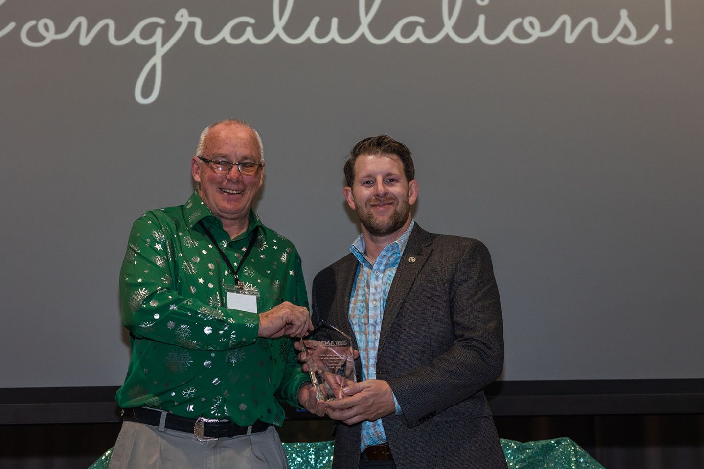 Two men on stage, one in green shirt and the other in a suit, holding an award in front of a “Congratulations!” backdrop.