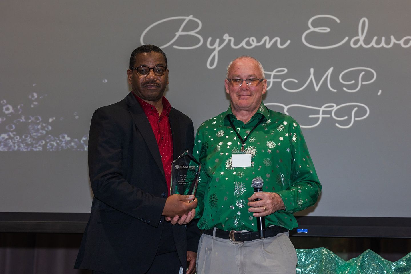 Two men on stage; one presents an award to the other. The awardee wears a green shirt, the presenter a suit.