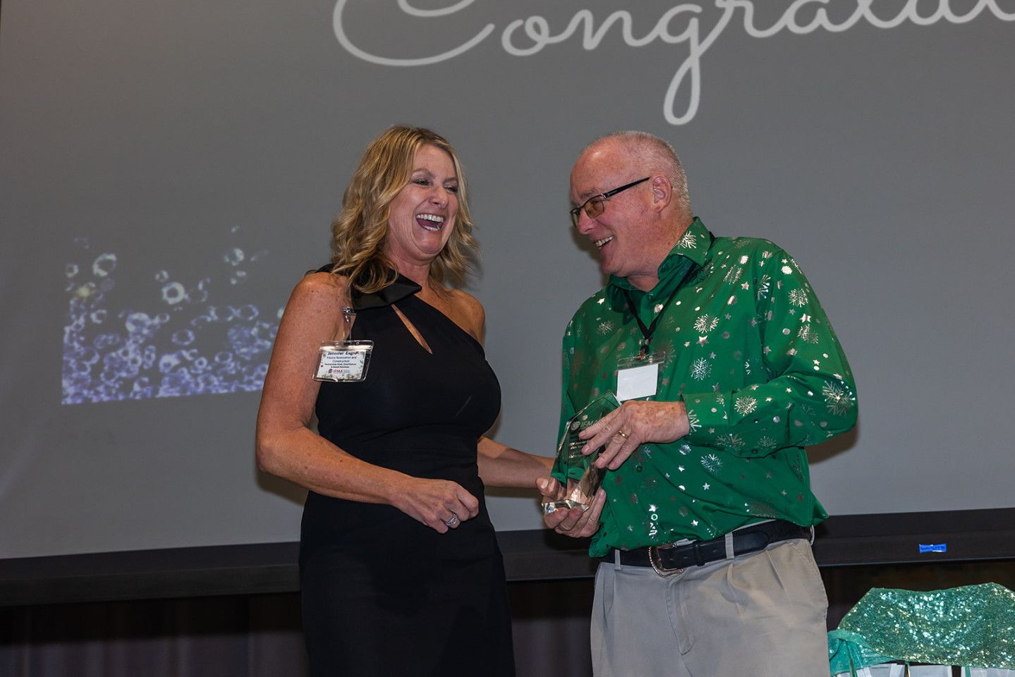 Woman in black dress laughing with man in green shirt, holding trophy on a stage.
