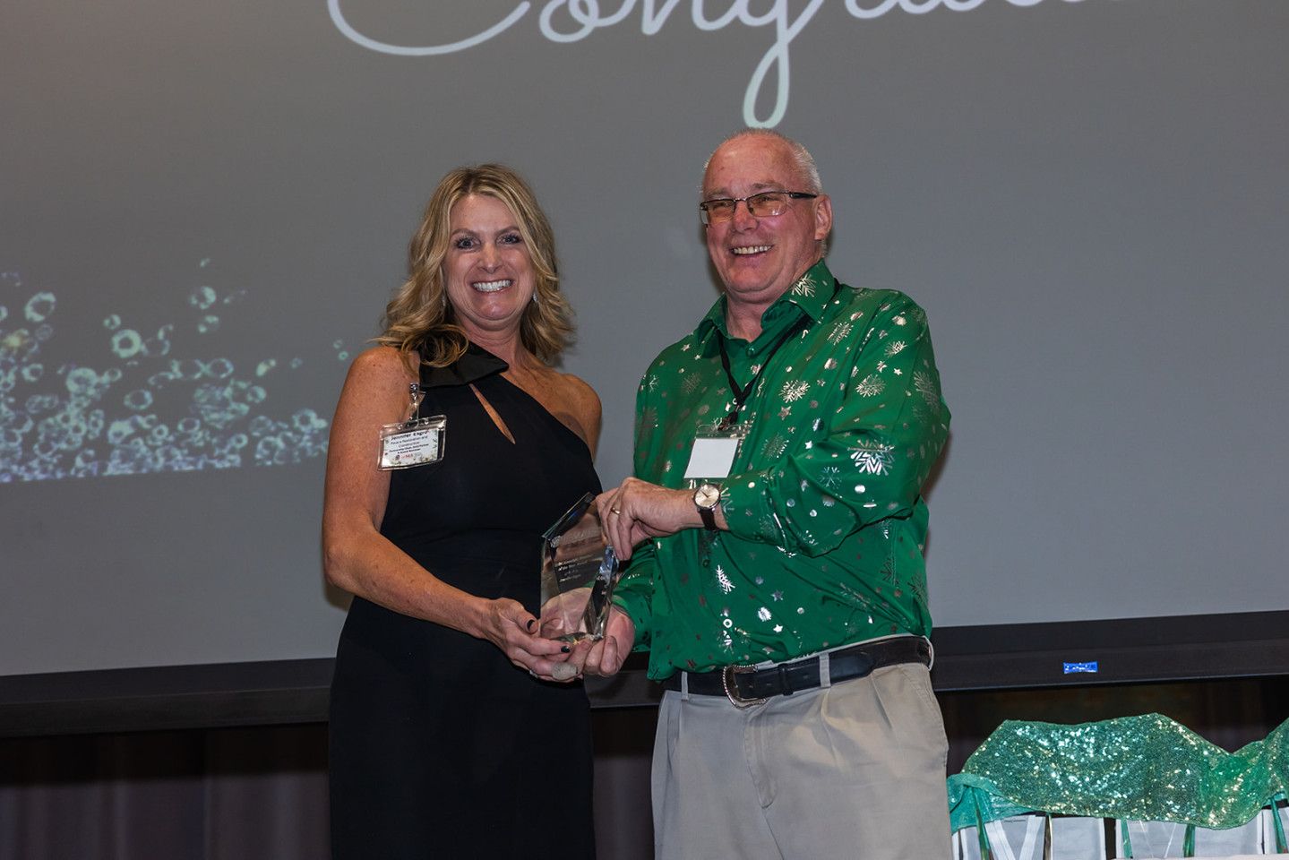 Woman in black dress presenting an award to a man in a green shirt. Stage backdrop with