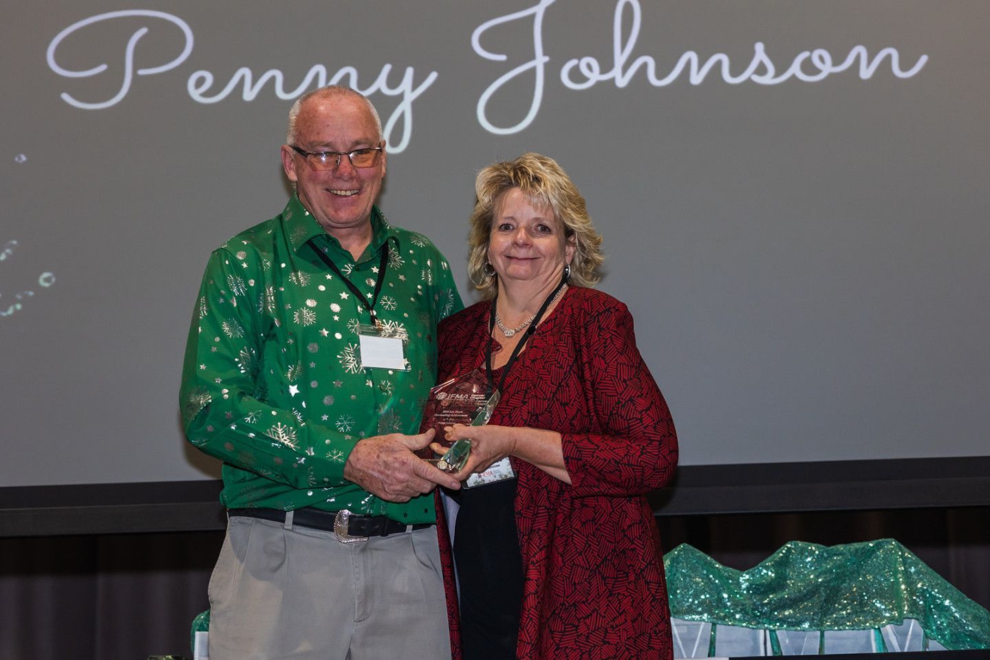 Man in green shirt presents an award to a woman in a red jacket, onstage under a name.