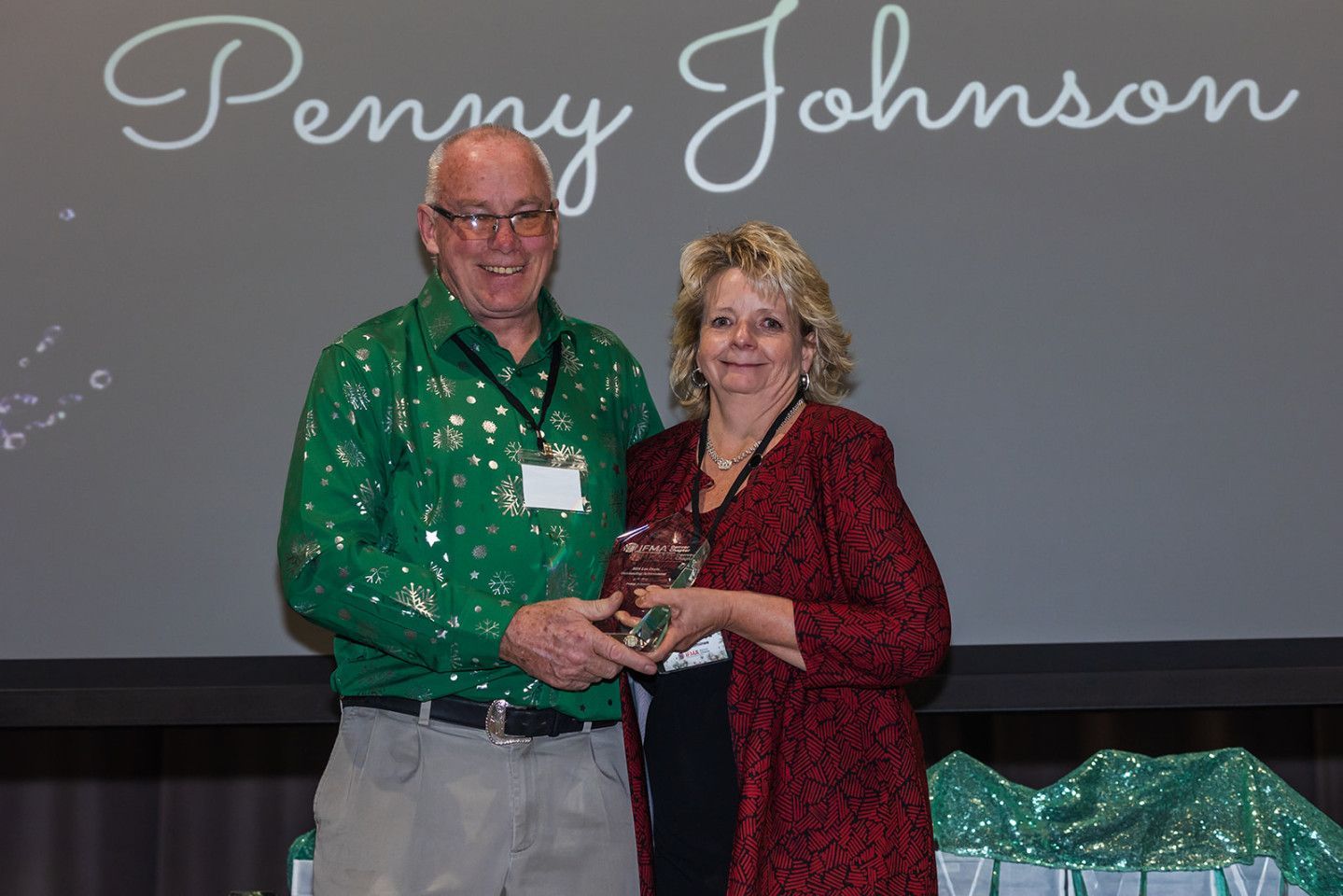 Man in green shirt presents an award to a woman in a red jacket. Penny Johnson is displayed.