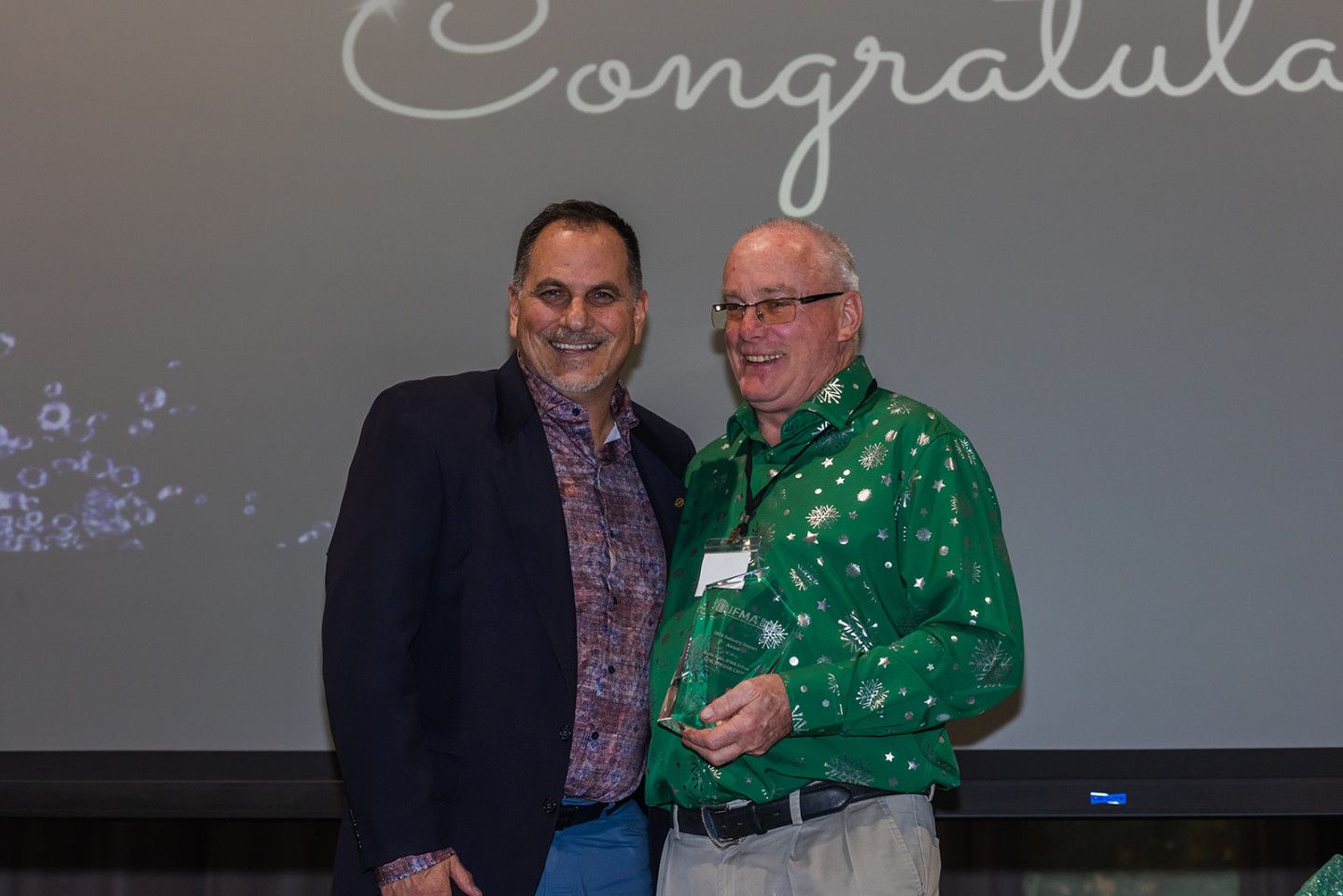 Two men stand together, one in green, holding an award. Grey screen reads