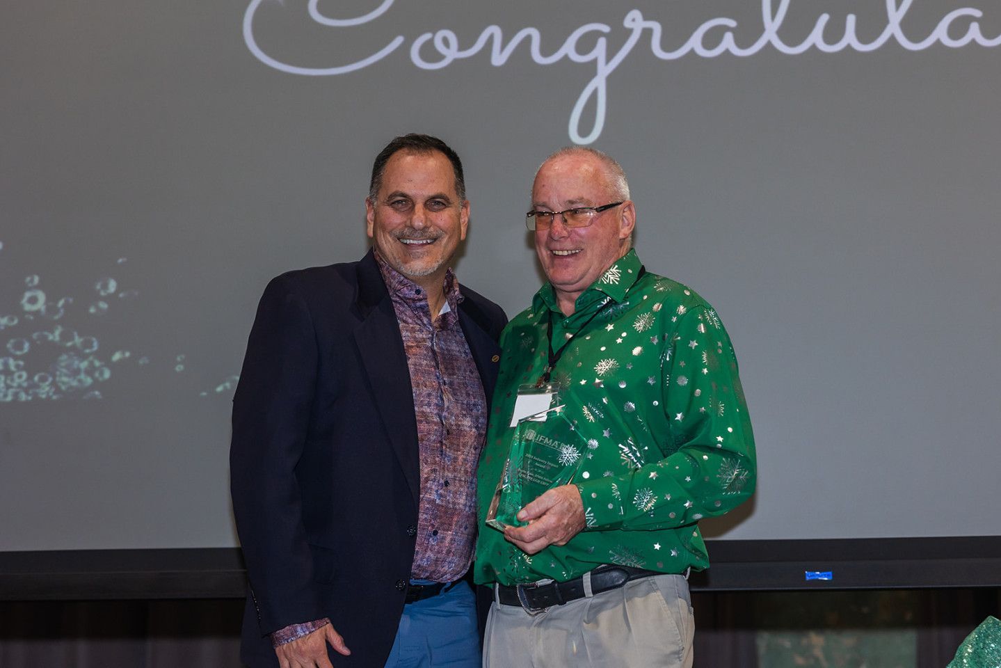 Two men smiling, one holding an award, in front of a screen that says