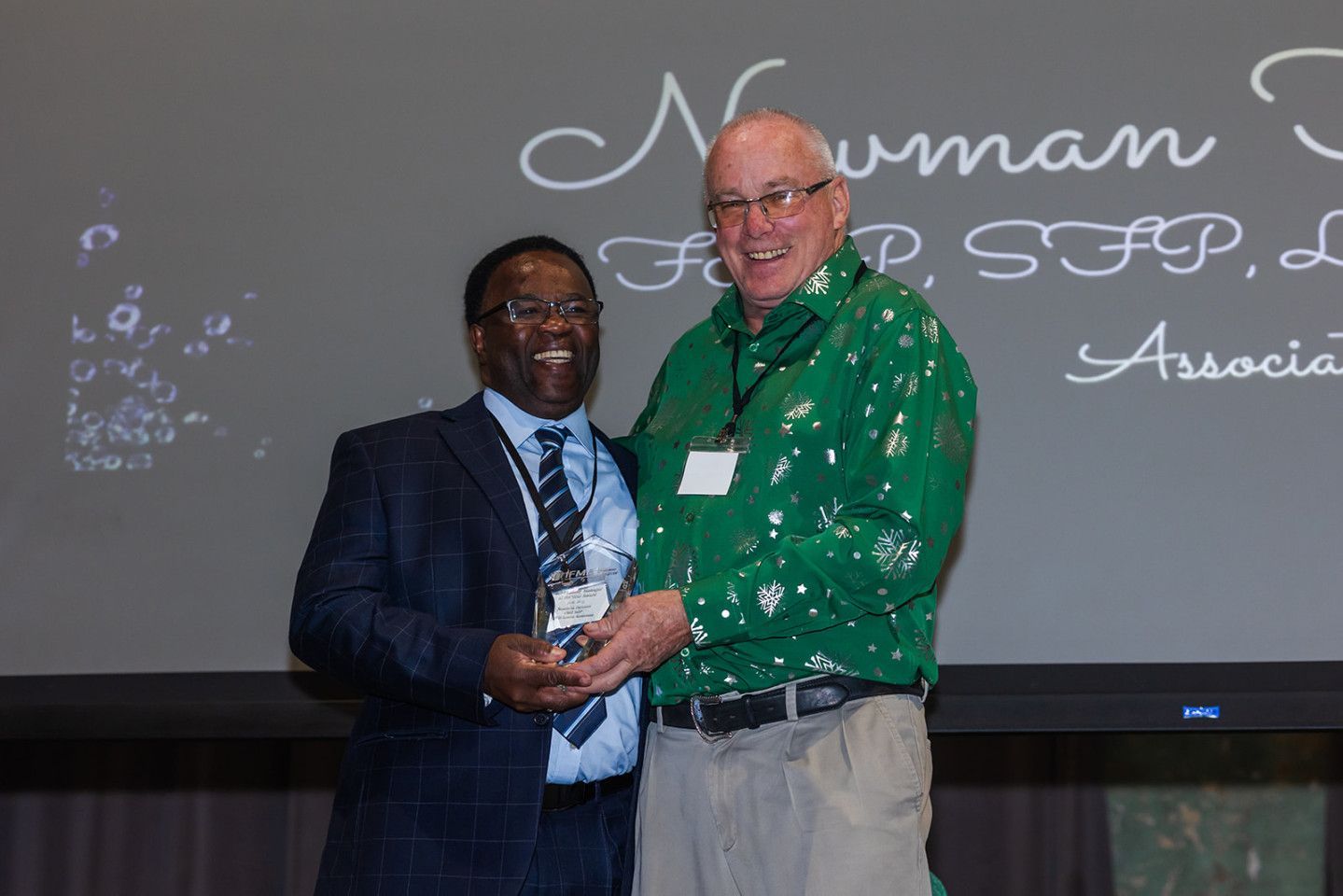 Two men on a stage. One in a suit gives an award to a man in a green shirt. A screen behind them has text.