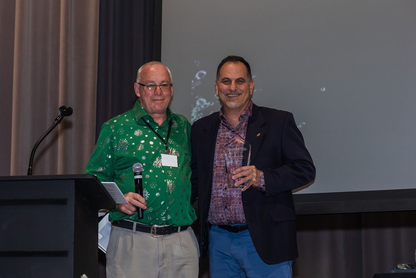Two men stand side-by-side, smiling, in front of a podium and screen.