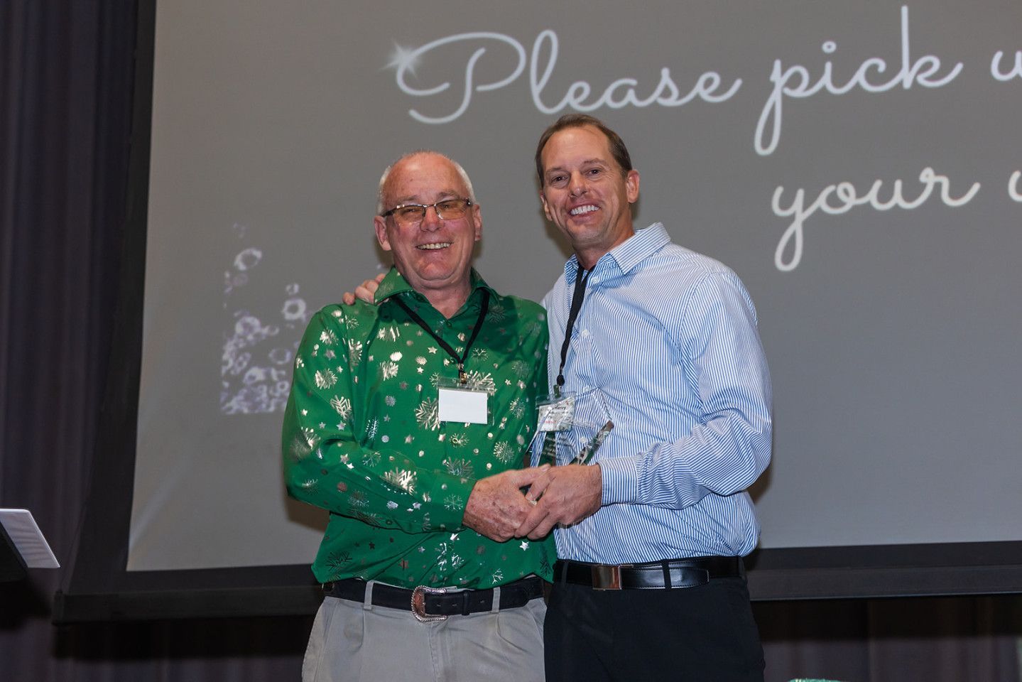 Two men holding an award on a stage. One in green shirt, the other in a blue patterned shirt.