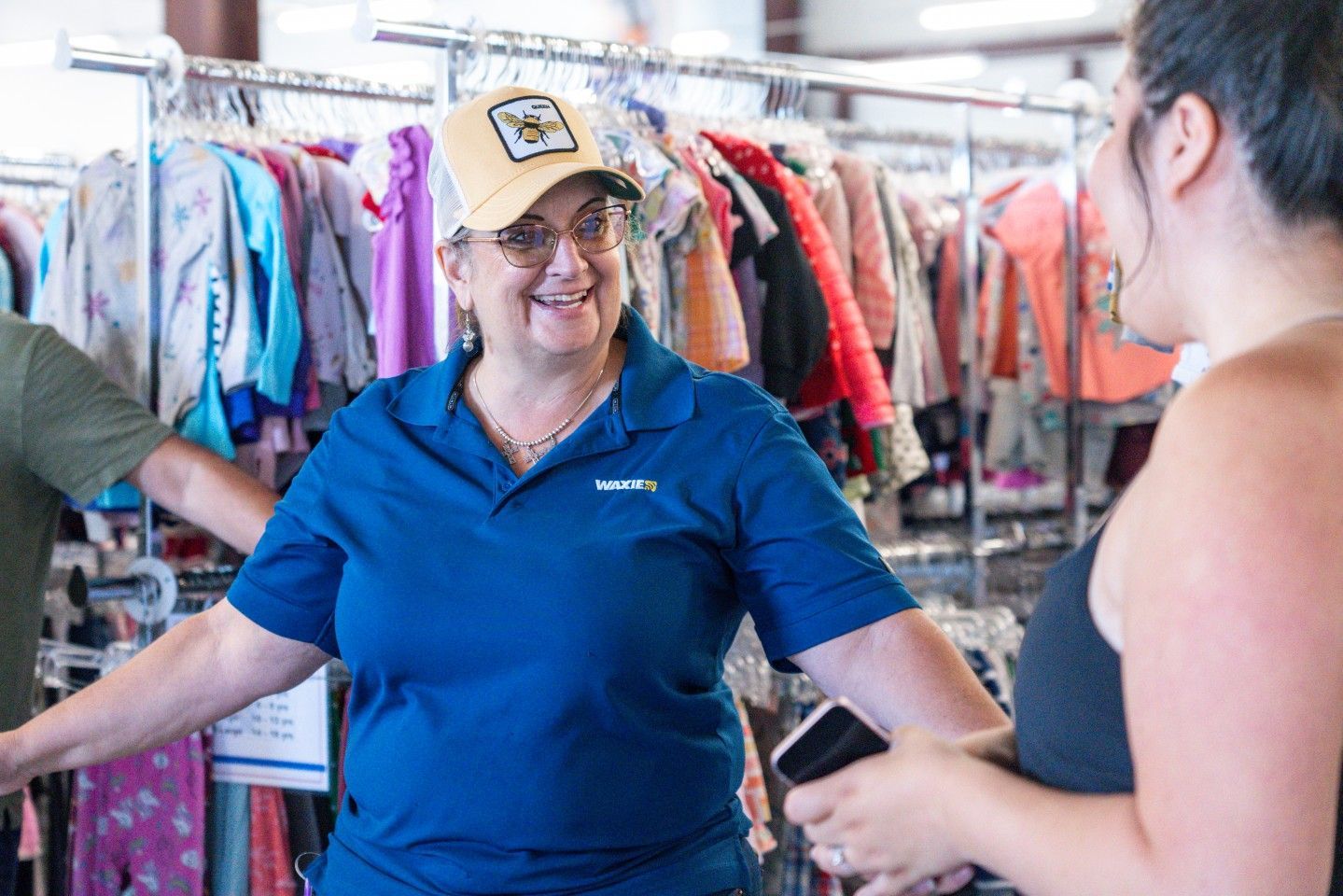 Woman in blue shirt smiles at another woman, in a thrift store, clothes racks visible.