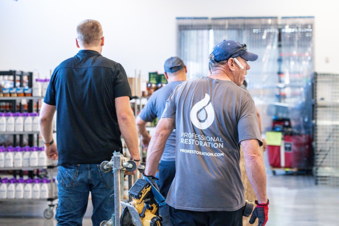 Three men walking with equipment in an interior space, one wearing a shirt with a company logo.