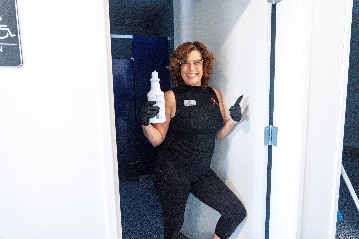 Woman in black, holding cleaning product, wearing gloves, in a restroom doorway, pointing.