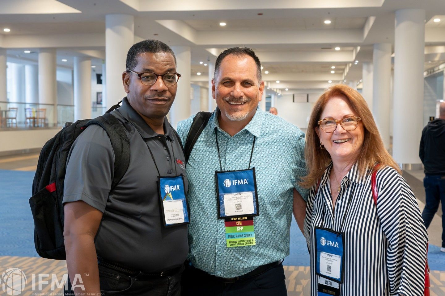 Three people smiling, posed together indoors with lanyards, appearing to be at a conference.