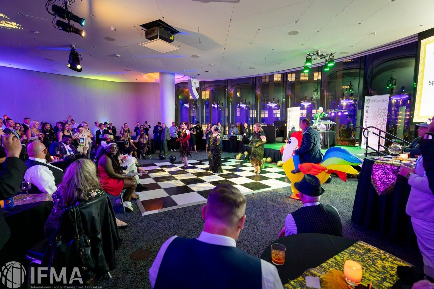 People at a gala event: checkered dance floor, stage with speakers, attendees seated at tables.