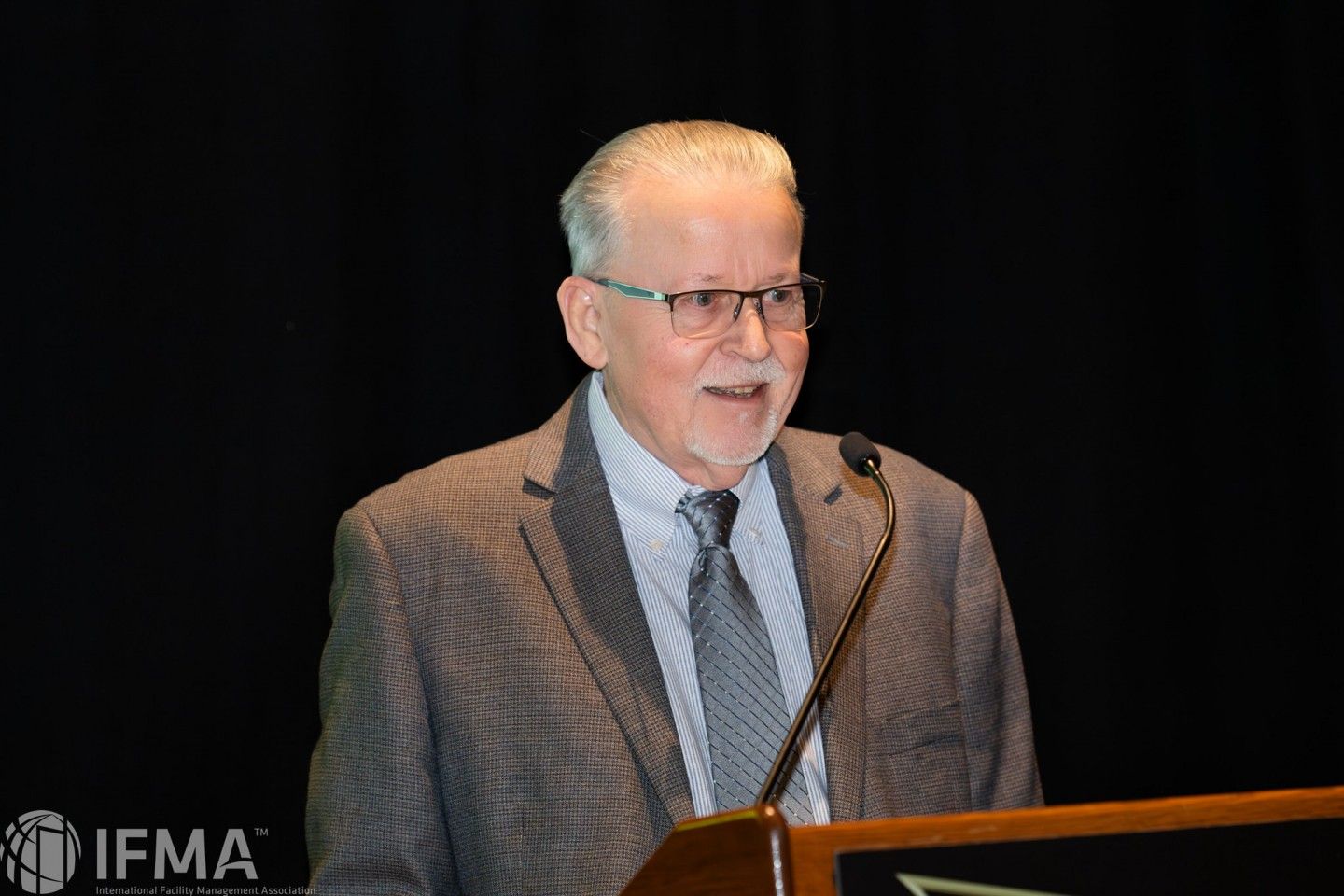 Man in glasses speaks at a podium with IFMA logo against a black backdrop.