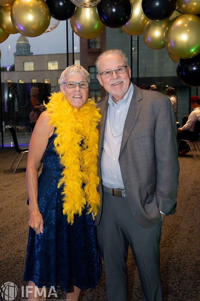 Two people smiling, standing together by gold and black balloons. Woman wears blue dress, yellow boa. Man in gray suit.