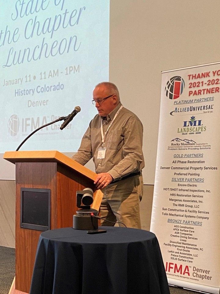Man speaking at a luncheon, standing at a podium. The sign reads 