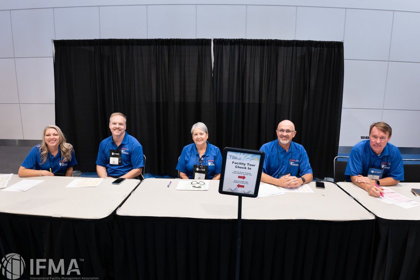 Five people in blue shirts sit behind a registration table with a black backdrop.