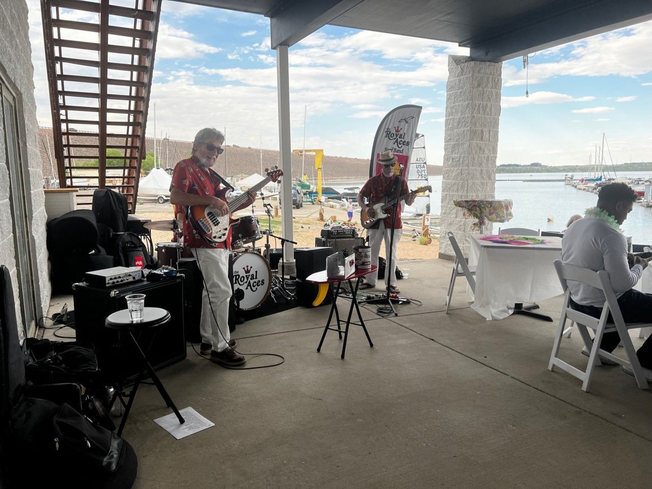 Band plays under a covered outdoor space near a harbor. Musicians wear Hawaiian shirts and play guitars.