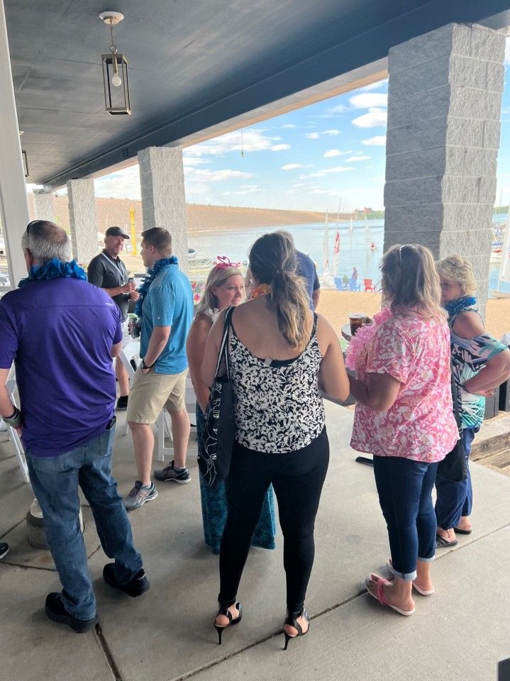 People gather on a patio overlooking a lake, wearing casual clothes and leis.