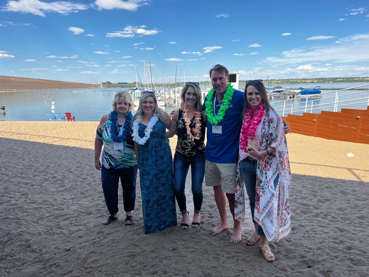 Five people with leis pose on a beach near a marina.