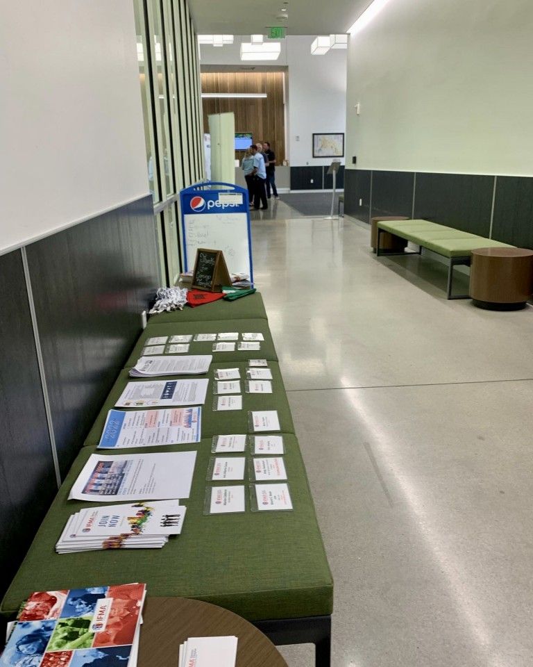 A table with brochures and name cards in a hallway with a bench, a soda machine, and a framed display.