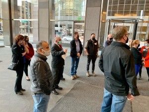 A group of people wearing masks stand in a building entrance with glass doors, looking toward the camera.