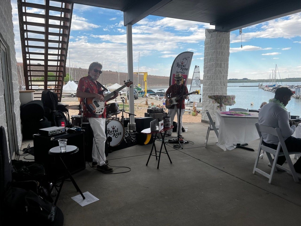 Band performing live music on a patio by a lake. Musicians play guitar, drums. Sky is cloudy.