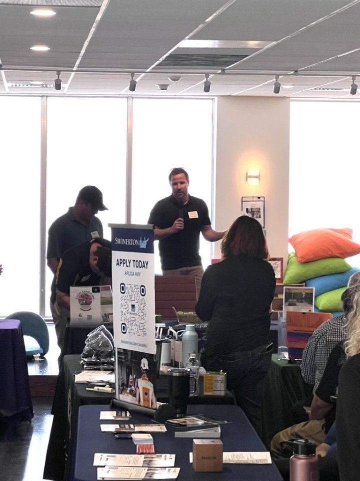 Man speaking at event; tables with products on display; people present; indoor setting.