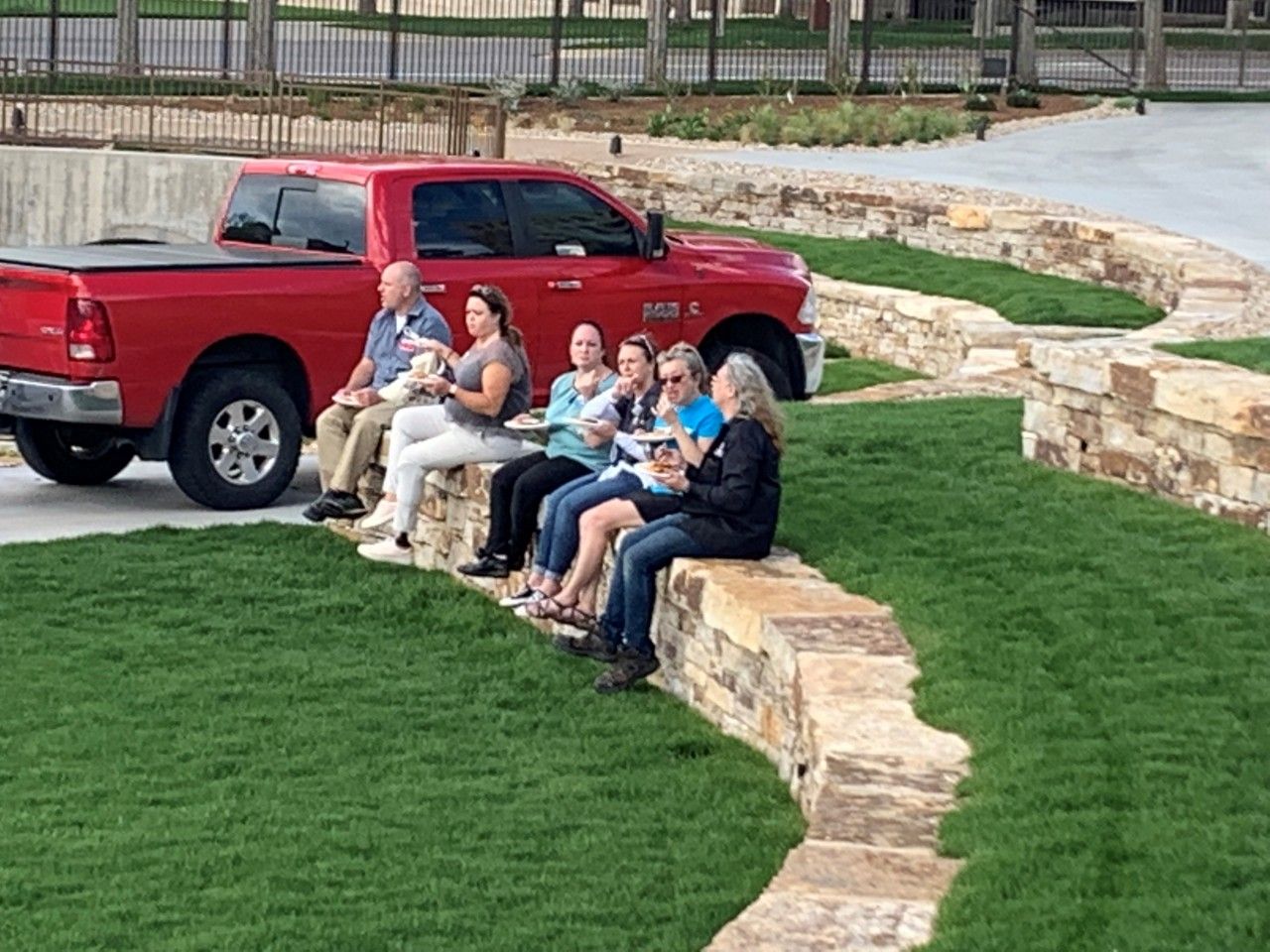 People sitting on a low stone wall by a grassy area and a red pickup truck.