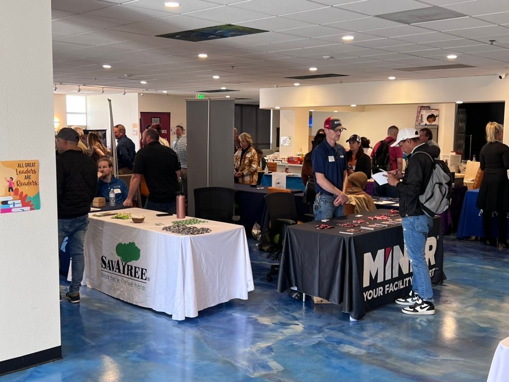 Event with booths and attendees. Tables with logos, people conversing in a large, open space with blue floor.