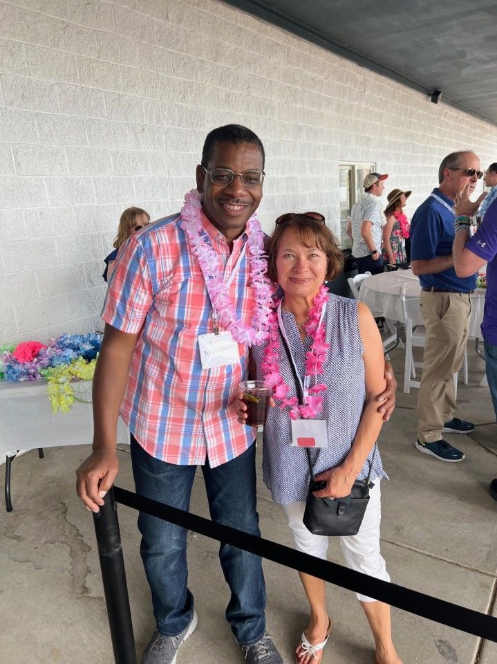 Man and woman pose for photo, both wearing leis, at outdoor event; man in checkered shirt, woman in blue and white.
