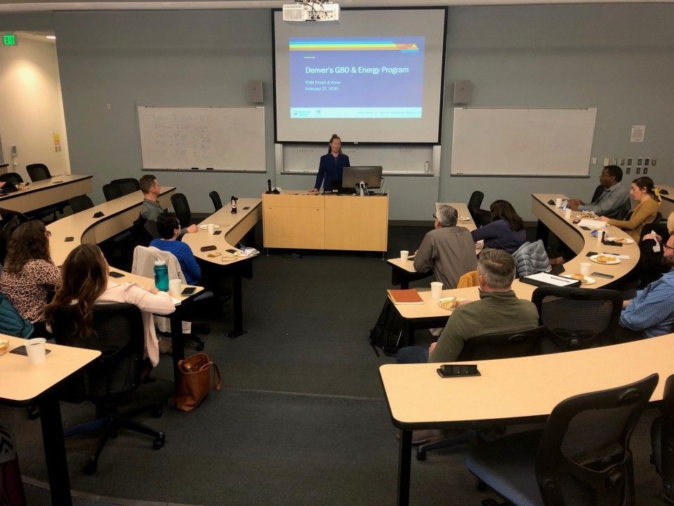 A woman presenting in a classroom with curved tables; audience members watch. A projector screen displays text.