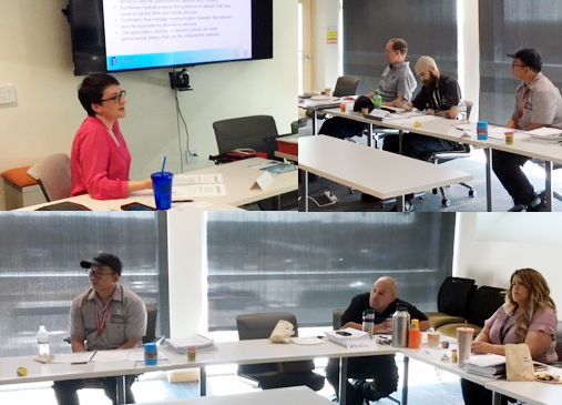 People in a meeting room, some looking at a screen, others at a table. A woman speaks at the front.