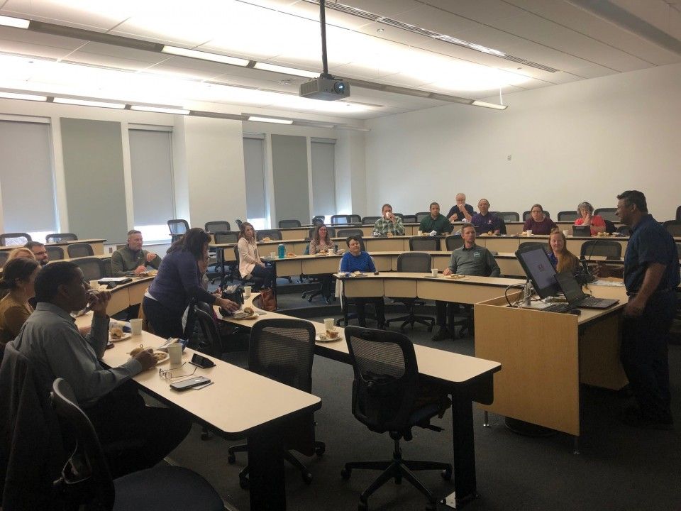 Classroom lecture: People seated at desks, one presenter at the front by a computer, all under bright lights.
