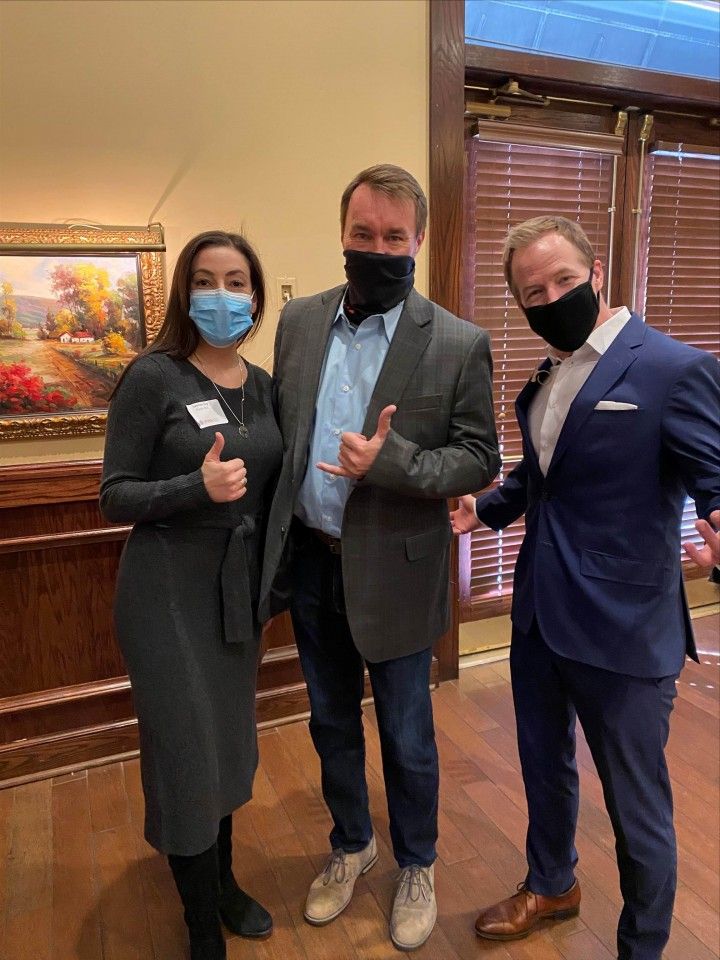 Three people wearing masks pose indoors; two giving thumbs-up, one making a hand gesture. Artwork and wood paneling visible.