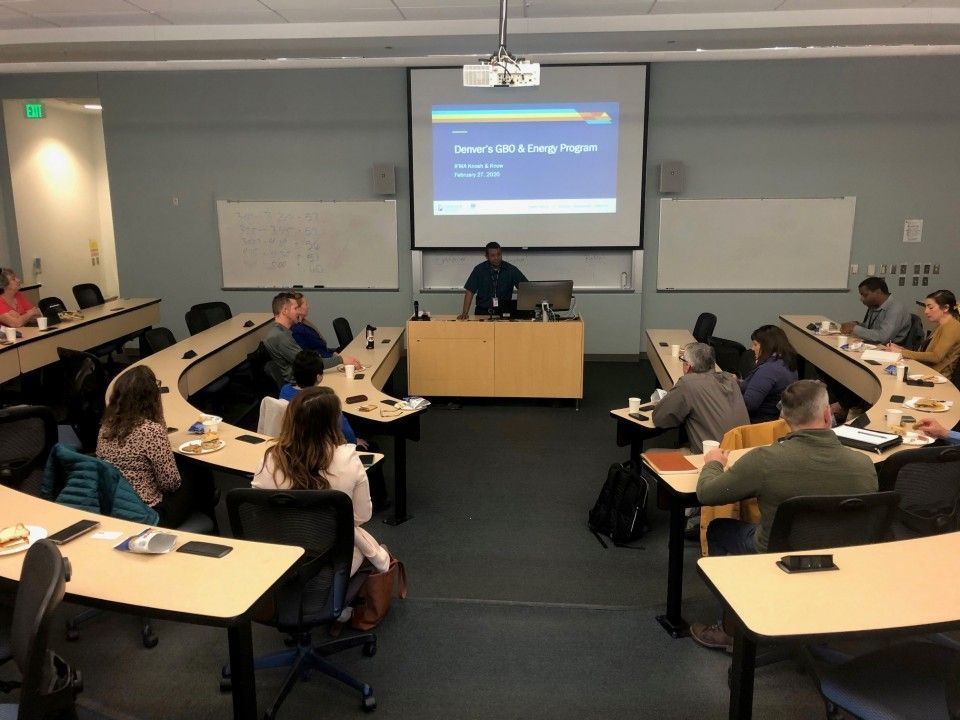 A man lectures in a classroom with a U-shaped table arrangement and people seated. A projector screen displays text.