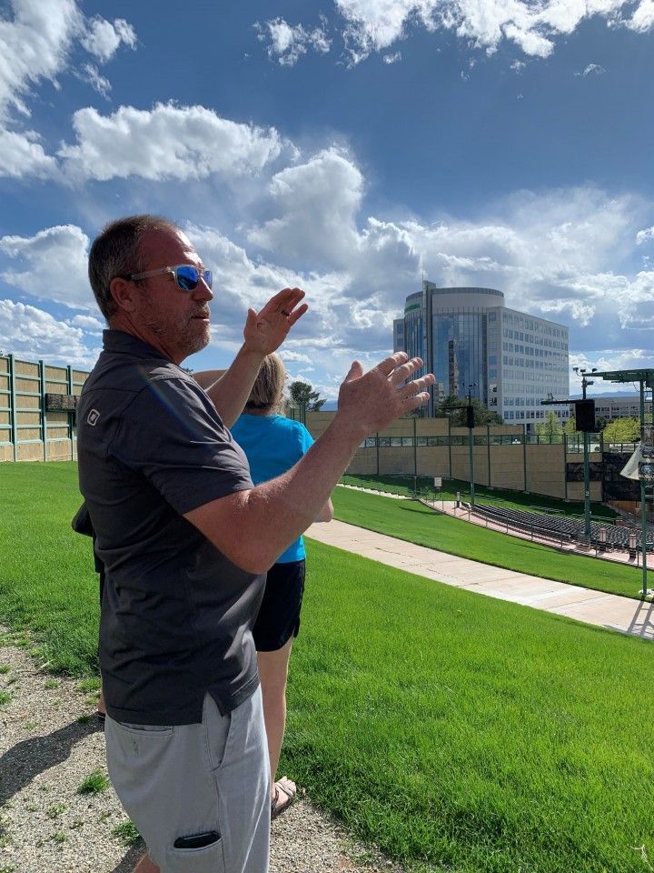 Man gesturing with hands outdoors, sunny day. Green grass slope, tall building in the background.