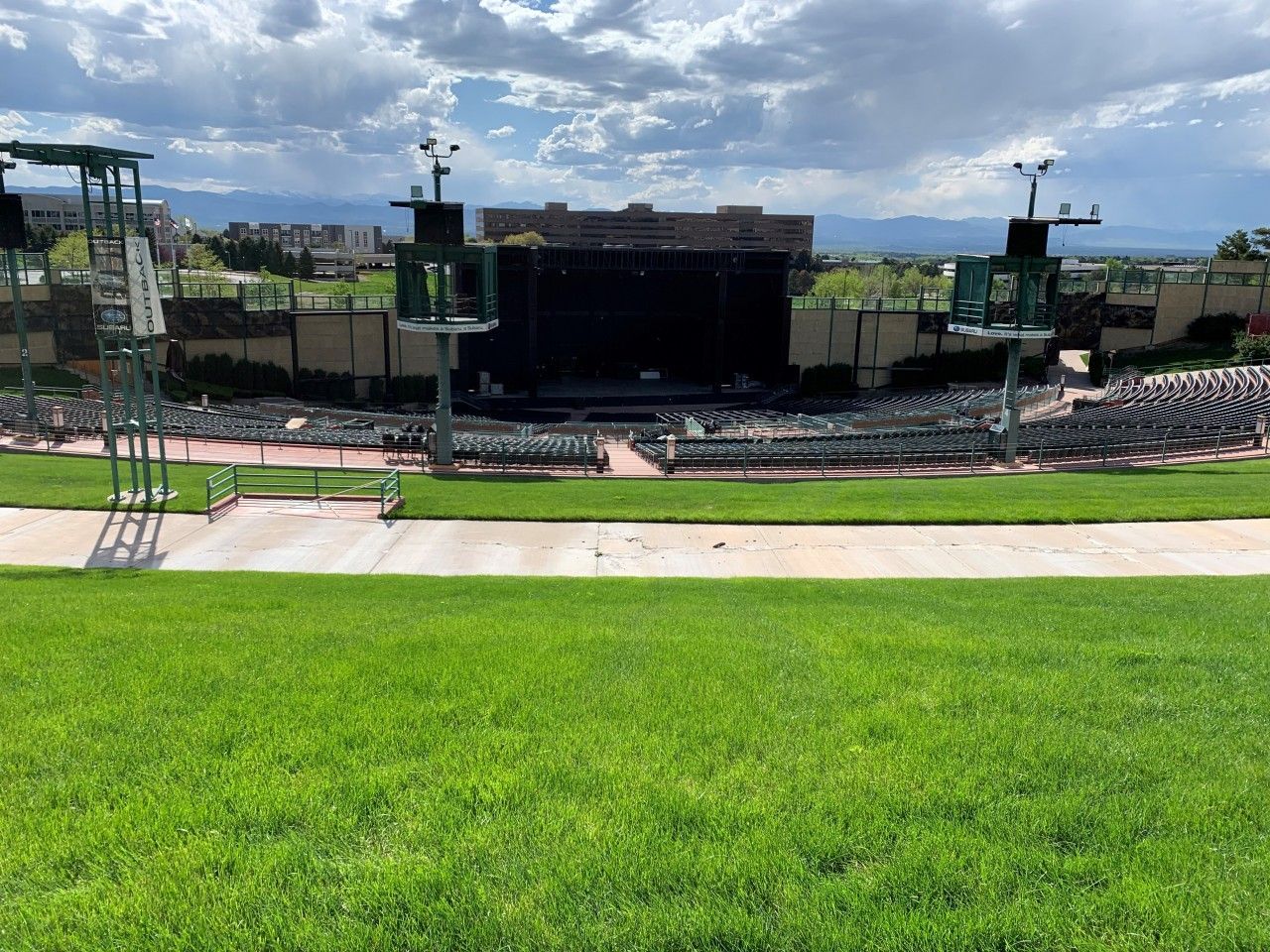 An outdoor amphitheater with stage, seating, and green lawn; mountains in background under cloudy sky.
