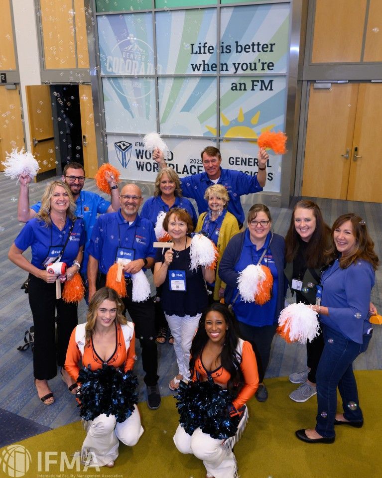 Group of people holding orange and white pom-poms, smiling. Banner behind them says “Life is better when you're an FM.”