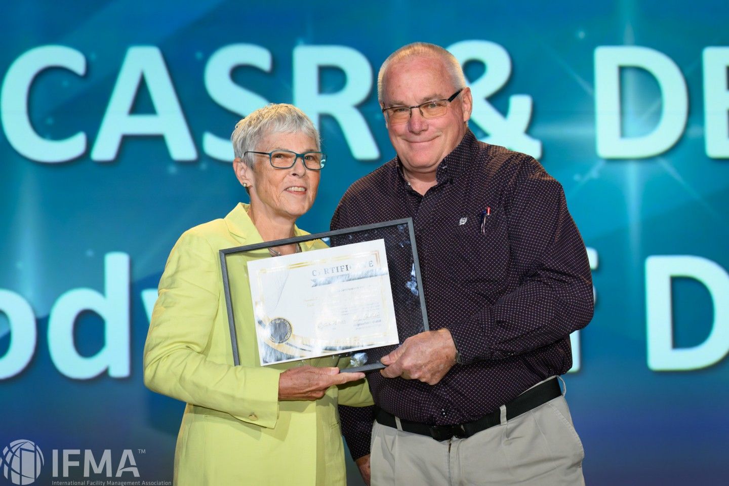 Woman in yellow jacket presents an award to a man in a patterned shirt on a stage, blue backdrop with text.